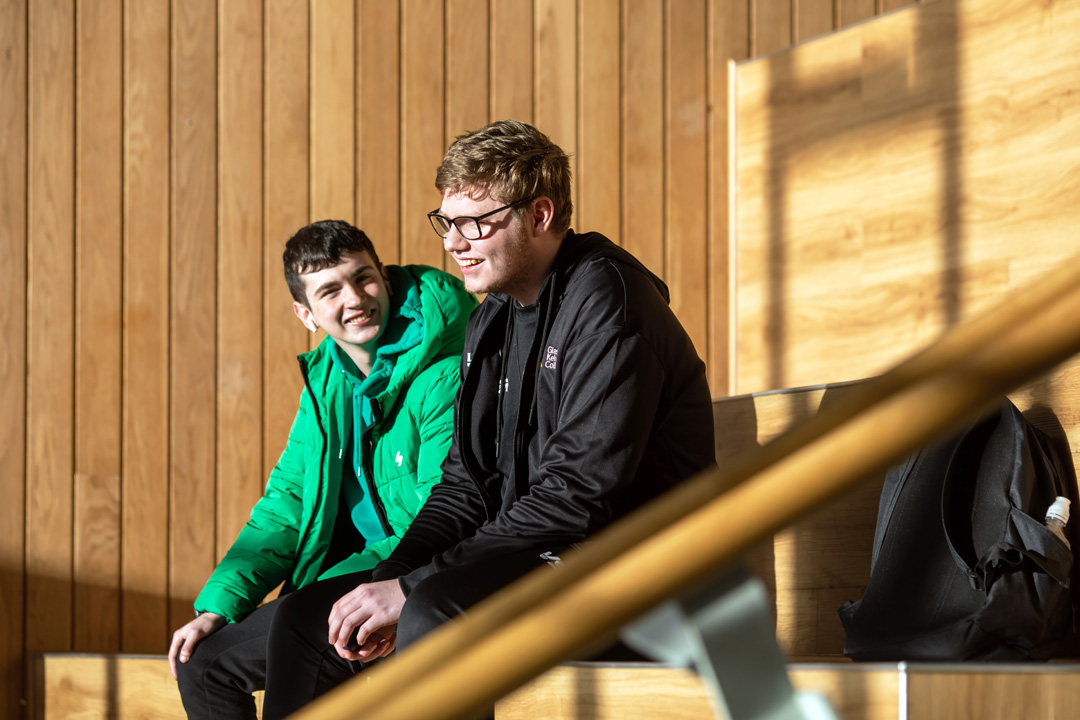 Two students sitting on tiered wooden seating at Springburn campus, chatting and enjoying the sunlight.