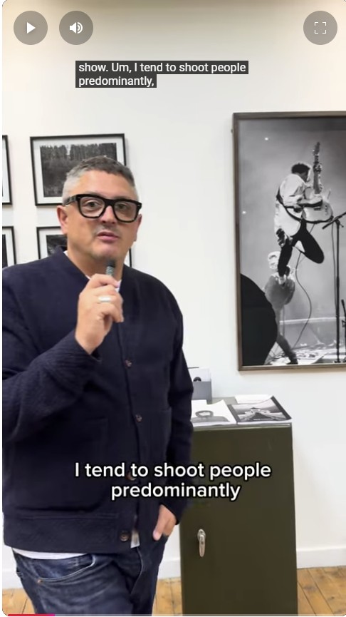 Photographer Mark Cameron speaking at an exhibition, standing in front of a wall of framed black and white portraits, including an action shot of a guitarist.
