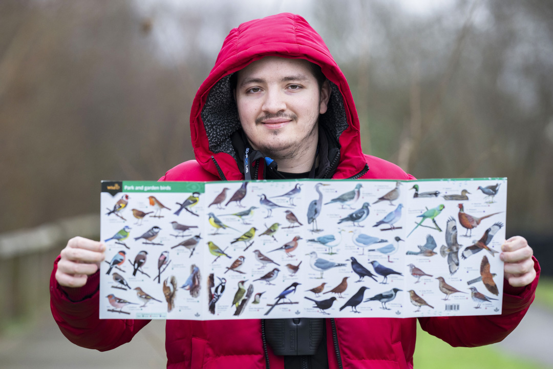 Smiling man in a red jacket holding a bird identification poster with various bird species displayed. Smiling man in a red jacket holding a bird identification poster with various bird species displayed.