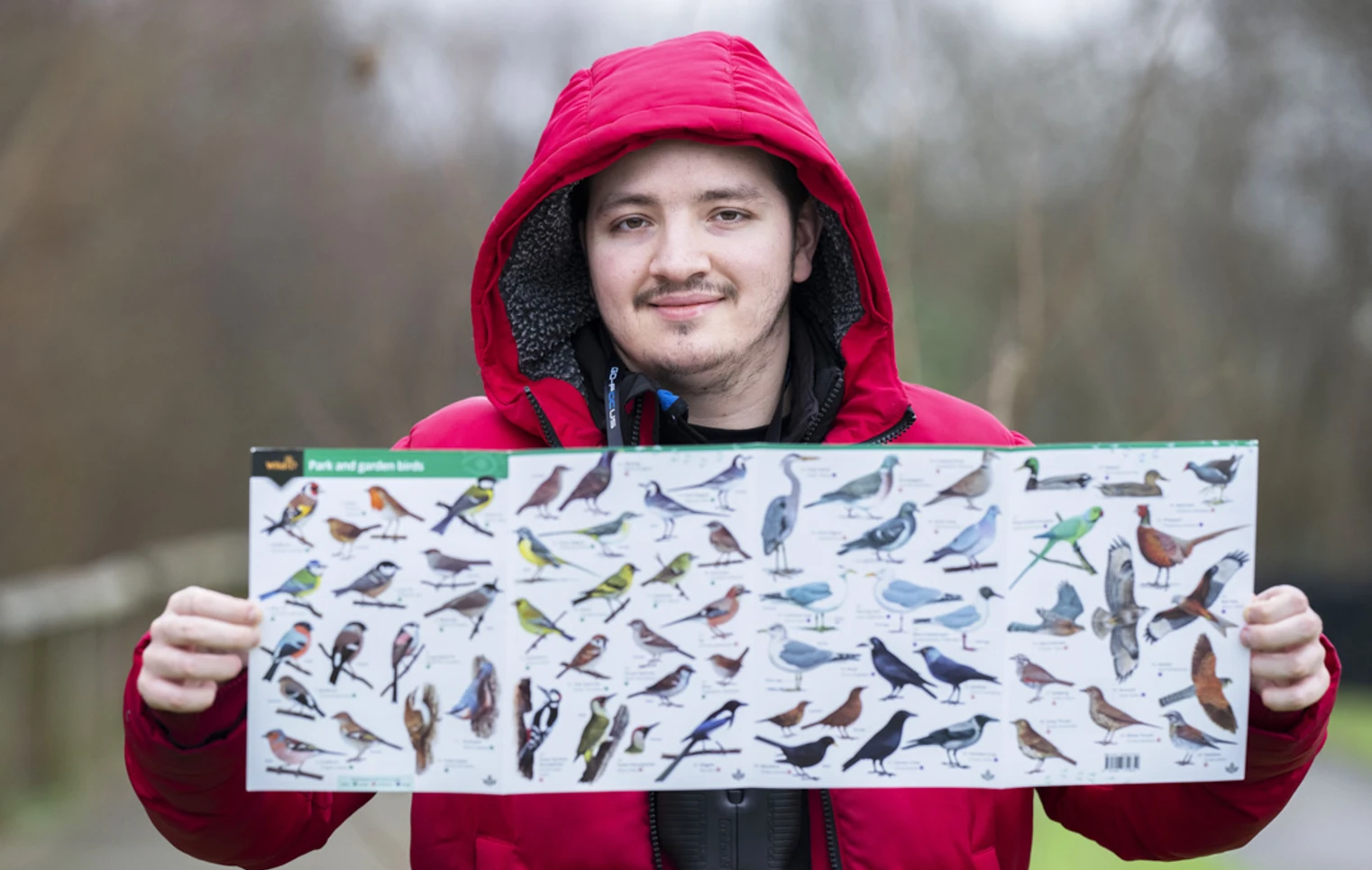 Smiling man in a red jacket holding a bird identification poster with various bird species displayed. Smiling man in a red jacket holding a bird identification poster with various bird species displayed.