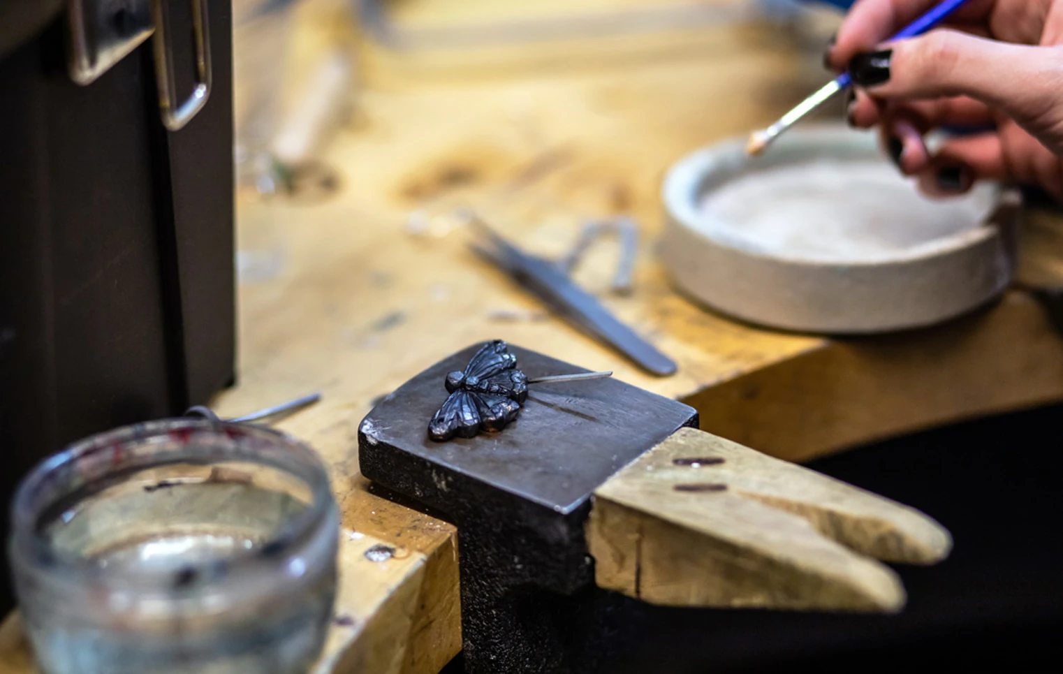 A close-up of a dark metal butterfly-shaped jewellery piece on a metal block, with an artist’s hand holding a fine brush to apply detail work. A close-up of a dark metal butterfly-shaped jewellery piece on a metal block, with an artist’s hand holding a fine brush to apply detail work.