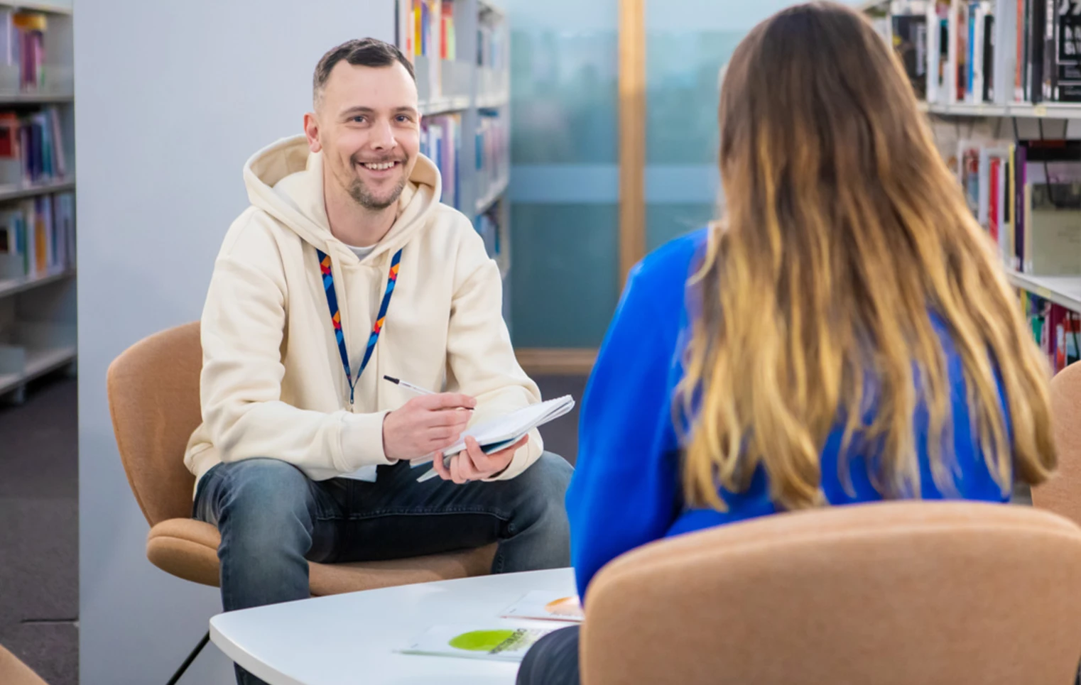 Glasgow Kelvin College staff member in a cream hoodie speaking with a student in a library setting, both seated and engaged in conversation with a notepad. Glasgow Kelvin College staff member in a cream hoodie speaking with a student in a library setting, both seated and engaged in conversation with a notepad.