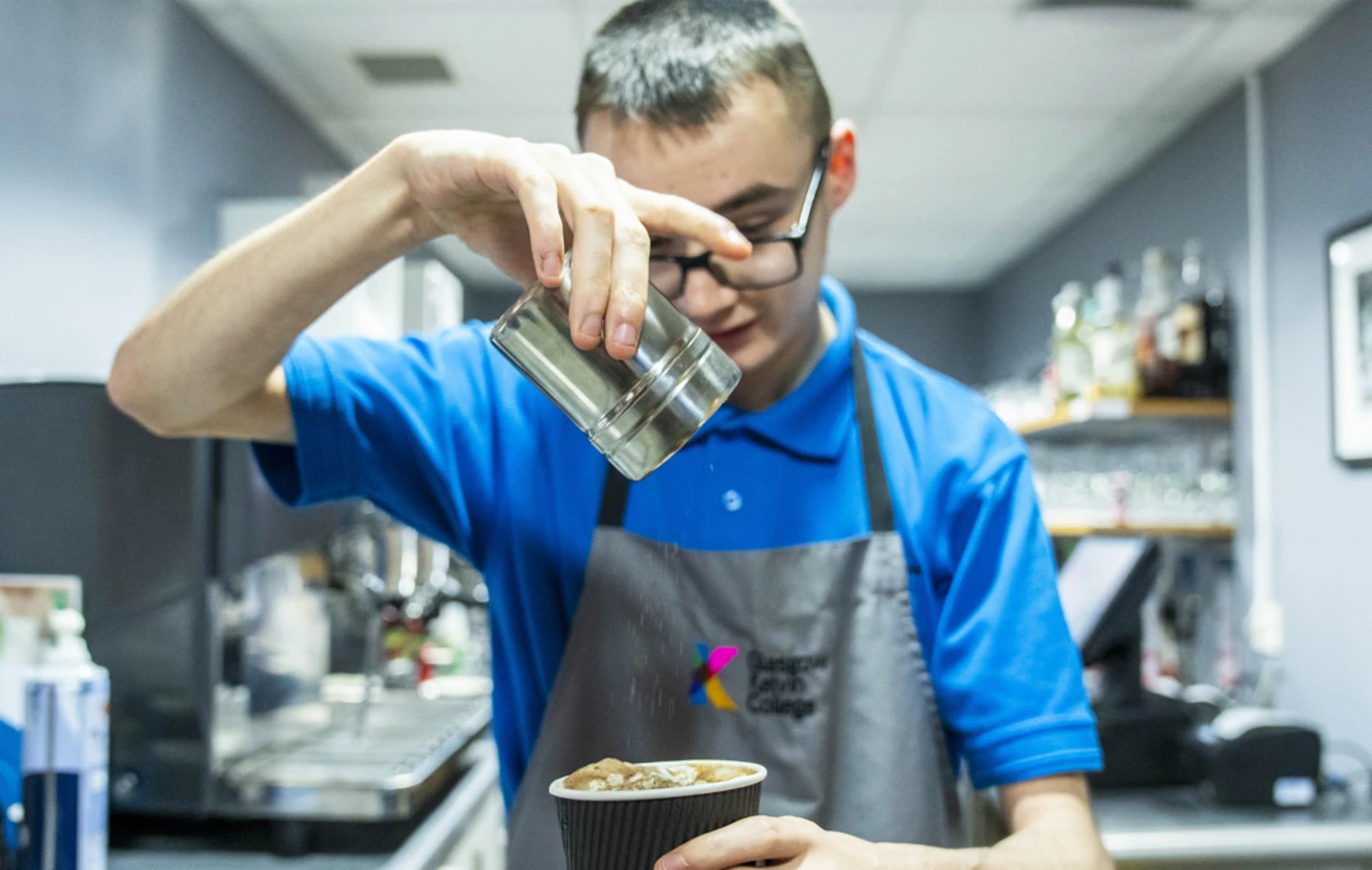 A young barista in a blue uniform sprinkles cocoa powder over a freshly prepared cup of coffee in a café setting. A young barista in a blue uniform sprinkles cocoa powder over a freshly prepared cup of coffee in a café setting.