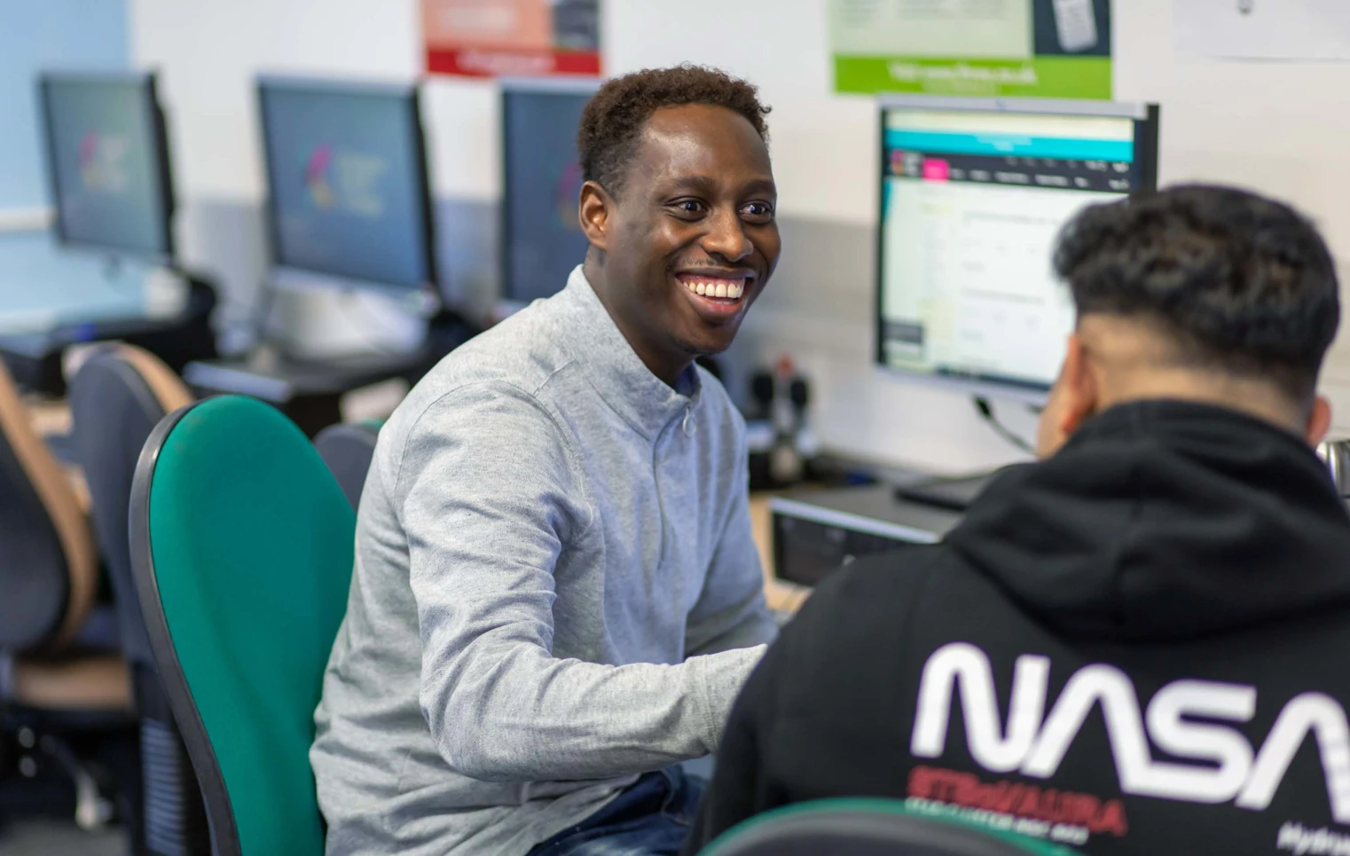 Two students working together at a computer in a business and administration course, one smiling while engaged in discussion. Two students working together at a computer in a business and administration course, one smiling while engaged in discussion.