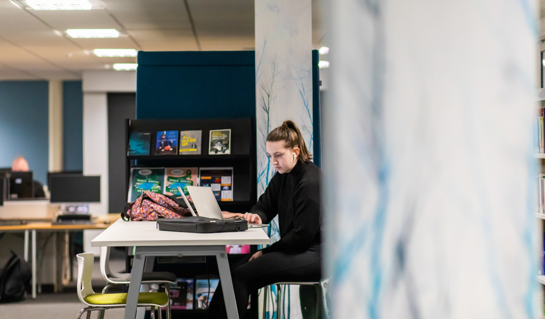 Student working on a laptop at a study desk in the Library and Flexible Learning Centre at Glasgow Kelvin College’s Easterhouse Campus, with bookshelves and resources in the background.