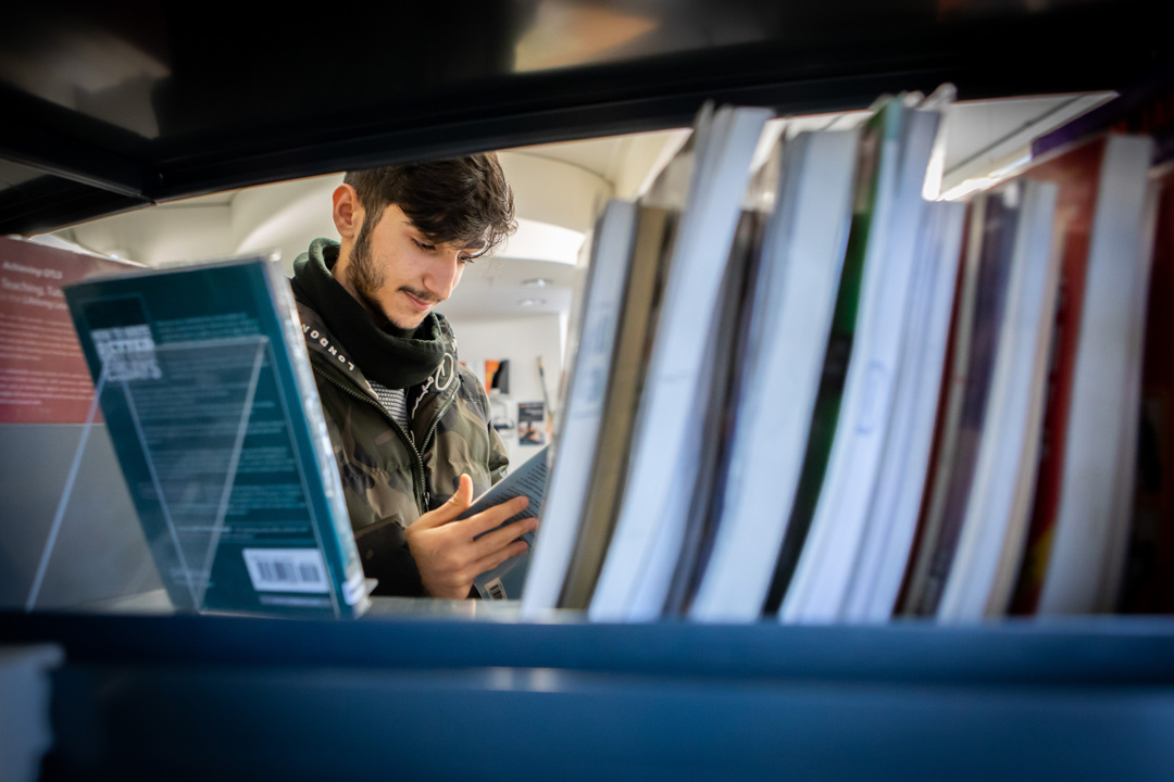 Student browsing and reading a book between shelves in the East End Campus library at Glasgow Kelvin College.