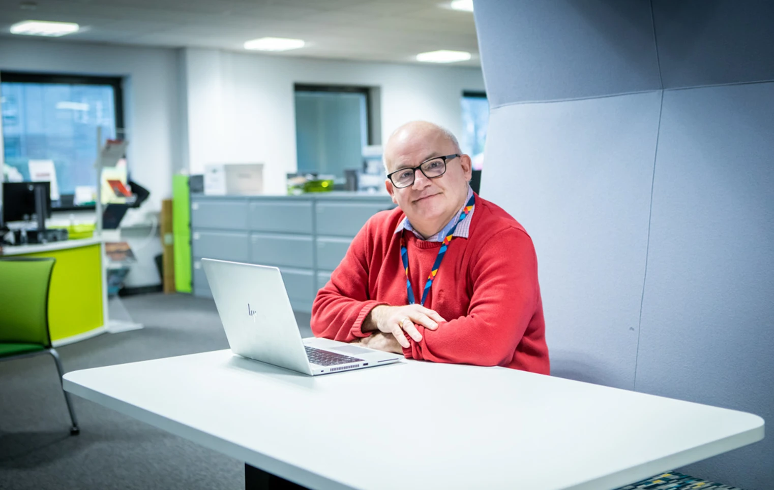 Smiling staff member in a red jumper sitting at a desk with a laptop in a modern office setting at Glasgow Kelvin College Smiling staff member in a red jumper sitting at a desk with a laptop in a modern office setting at Glasgow Kelvin College
