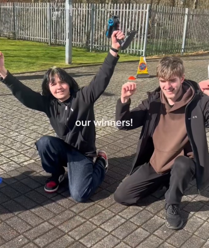 Two students celebrating after winning the Glasgow Kelvin College CAD car race, holding remote controls outdoors.