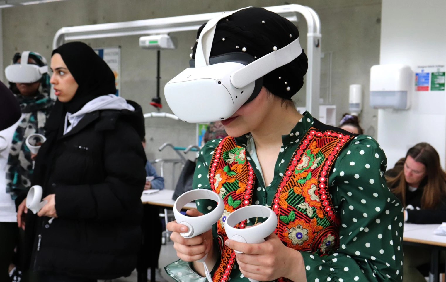 A woman in a green polka-dot dress with a colorful embroidered vest wears a VR headset and holds controllers while engaging in a virtual training session, surrounded by other students. A woman in a green polka-dot dress with a colorful embroidered vest wears a VR headset and holds controllers while engaging in a virtual training session, surrounded by other students.