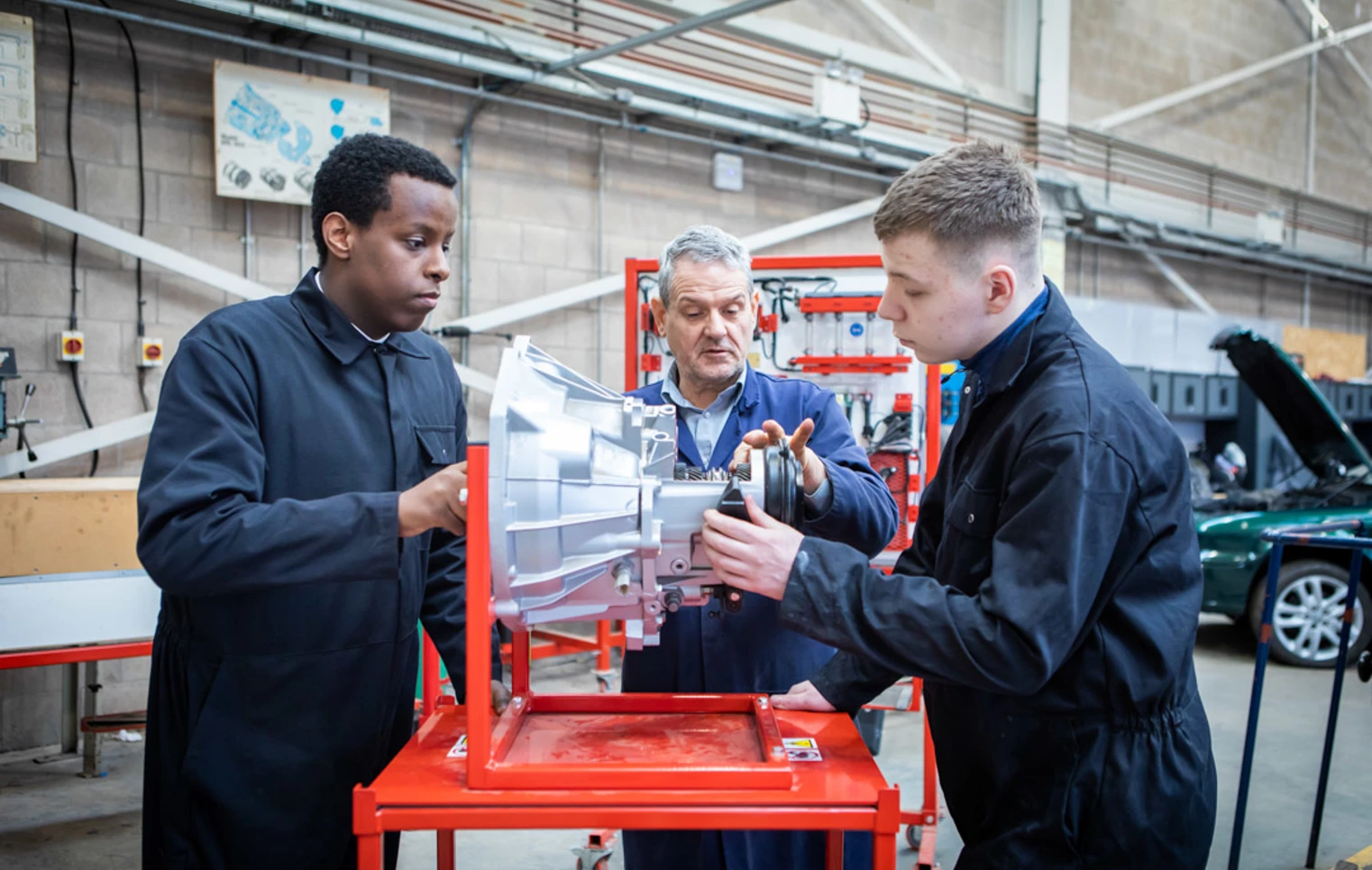 Automotive instructor guiding two students in overalls as they work on a mechanical component in a workshop. Automotive instructor guiding two students in overalls as they work on a mechanical component in a workshop.