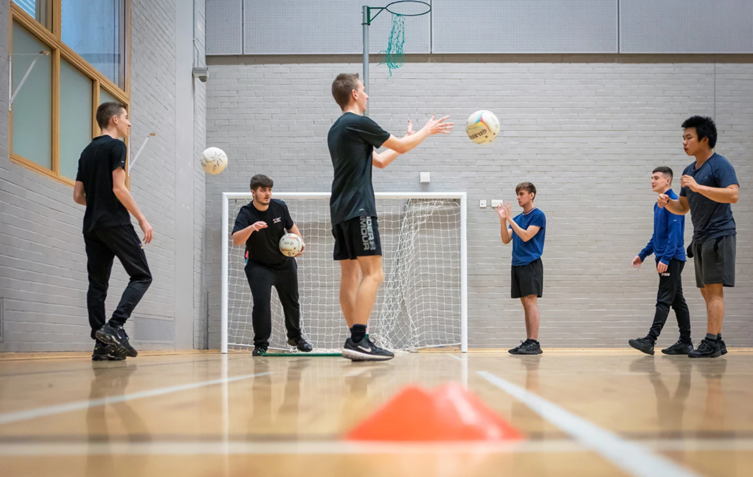 Students in a games hall tossing netballs in a circle as part of a group activity. Students in a games hall tossing netballs in a circle as part of a group activity.