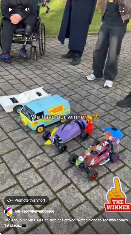 A group of people standing around three colorful, custom-built model cars at the finish line of the Annual Kelvin CAD Car Race. The winning car, a bright blue and yellow van, is in the foreground with a "Winner" sticker, while the other two cars, one purple and one red, are beside it on a paved surface.