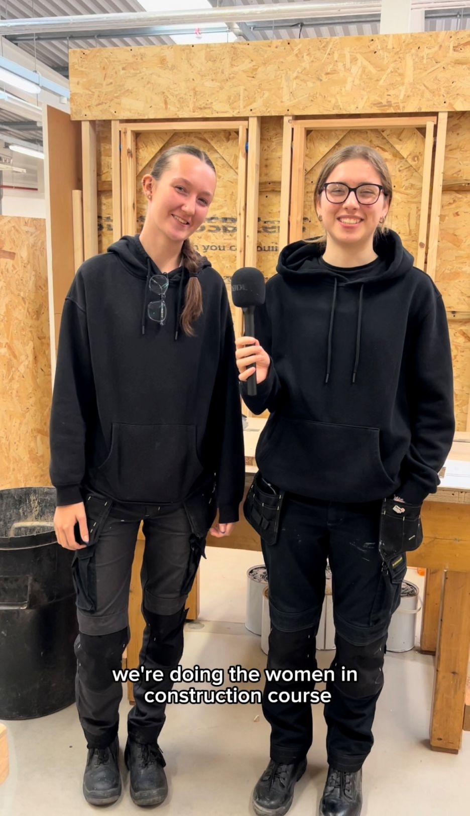 Two women construction students stand smiling in a workshop, both wearing black hoodies and work trousers. One holds a microphone as they talk about the Women in Construction course.