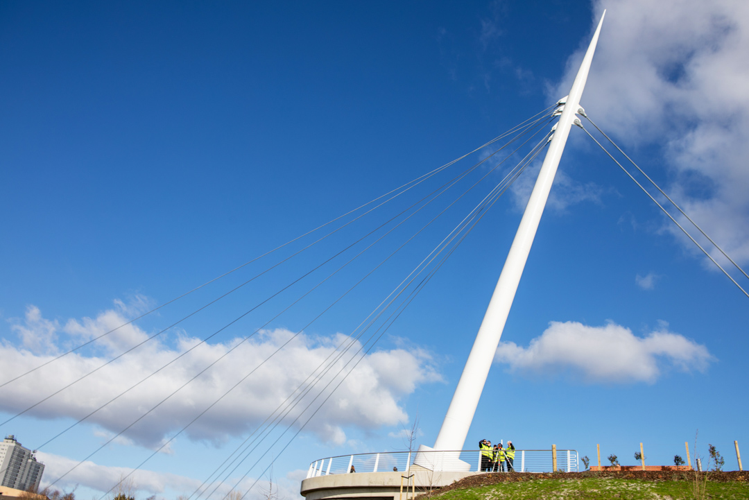 A group of civil engineering students and a lecturer in high-visibility vests conducting a land surveying exercise on the Stockingfield Bridge, viewed from a lower angle against the structure’s towering white support beam.