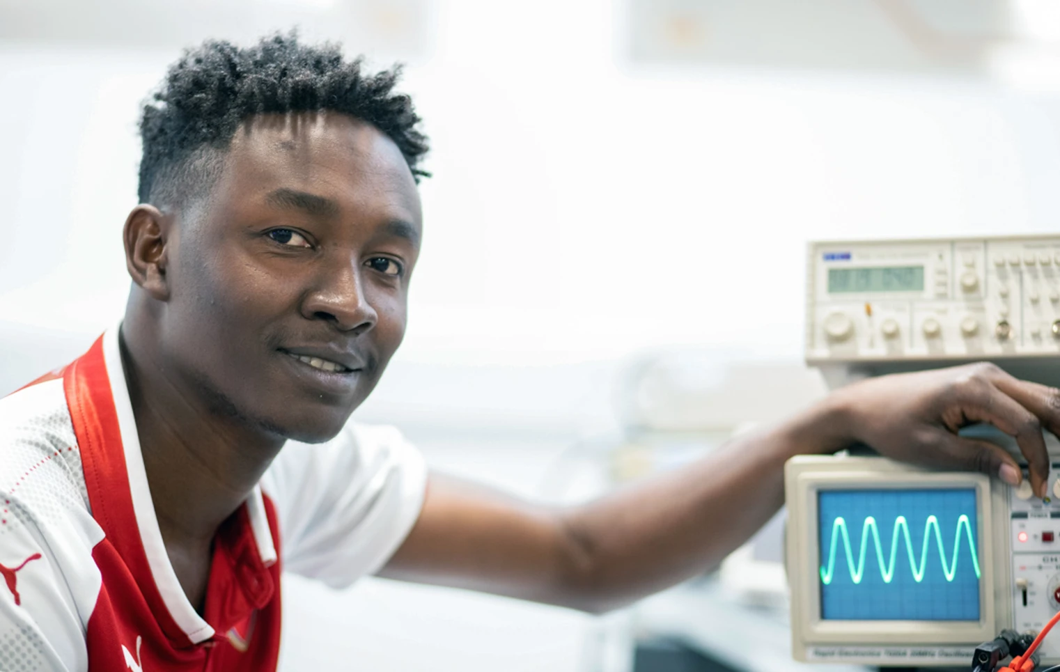 Close-up of a student in a red football jersey adjusting an oscilloscope, displaying a waveform on the screen. Close-up of a student in a red football jersey adjusting an oscilloscope, displaying a waveform on the screen.