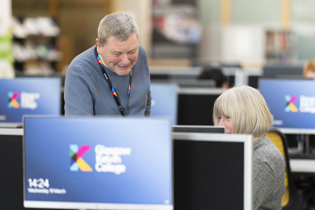 Staff member supporting a student in the computer suite at Glasgow Kelvin College, with college branding visible on screens.
