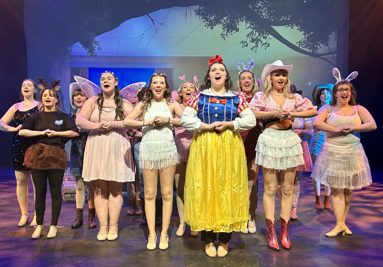Ensemble cast dressed in colorful costumes, singing joyfully on stage during a musical theatre production.